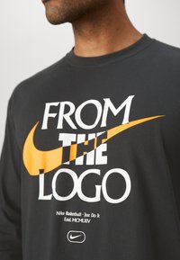 Black cotton t-shirt featuring bold white and orange text "FROM THE LOGO" with Nike Swoosh logo, crew neck design, and relaxed fit.