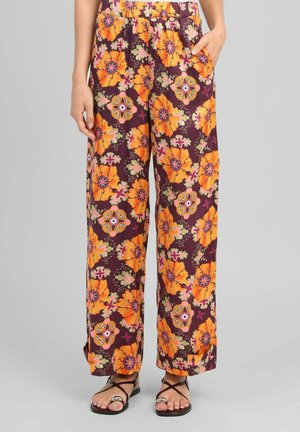 Femme portant un pantalon ample avec un grand imprimé floral orange sur fond violet foncé et des sandales noires à brides.