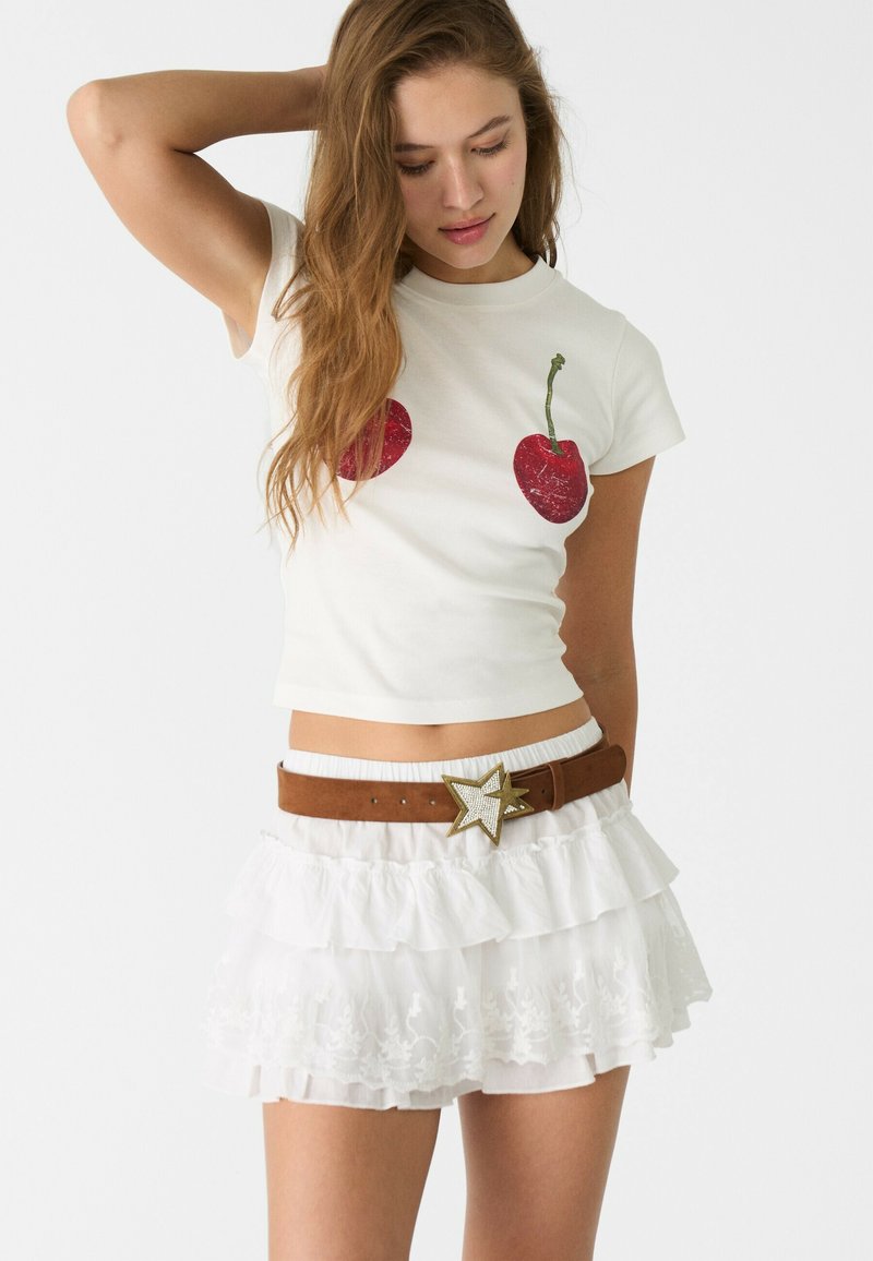 Witte cropped t-shirt met een rode kersengrafiek, gecombineerd met een witte minirok met ruffles en een bruine riem met een stervormige gesp.