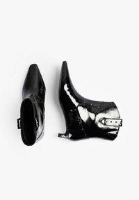 Botines de charol negro con puntera puntiaguda, tacón bajo, tachuelas decorativas y un detalle de correa en el tobillo. Textura de superficie lisa y brillante.
