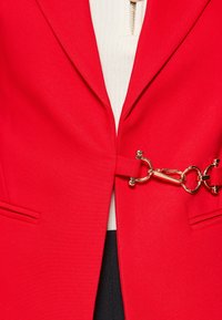 Rød blazer med et struktureret design, detaljer med guldfarvet hardware-lukning og sidelommer. Båret over en hvid ribbet top.