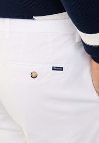 Pantalones de algodón blancos con un bolsillo trasero abotonado, que presentan una etiqueta azul marino y una cinturilla azul marino contrastante con rayas blancas.