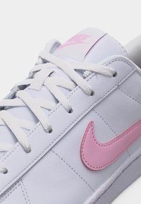 Λευκό αθλητικό παπούτσι Nike με ροζ λογότυπο swoosh, λευκά κορδόνια και ραφή στο δερμάτινο επάνω μέρος.