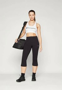 Svart kapad leggings och en vit crop top med "Champion"-text. Modellen håller en svart träningsväska och bär svarta sportskor. Släta texturer.