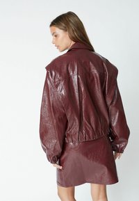 Giacca in finta pelle bordeaux con vestibilità rilassata, spalle basse e orlo elastico, indossata con una gonna coordinata in finta pelle.