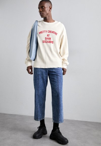 Beige ribbed sweatshirt with red text, paired with light blue wide-leg jeans. Black sneakers and a denim jacket slung over the shoulder.