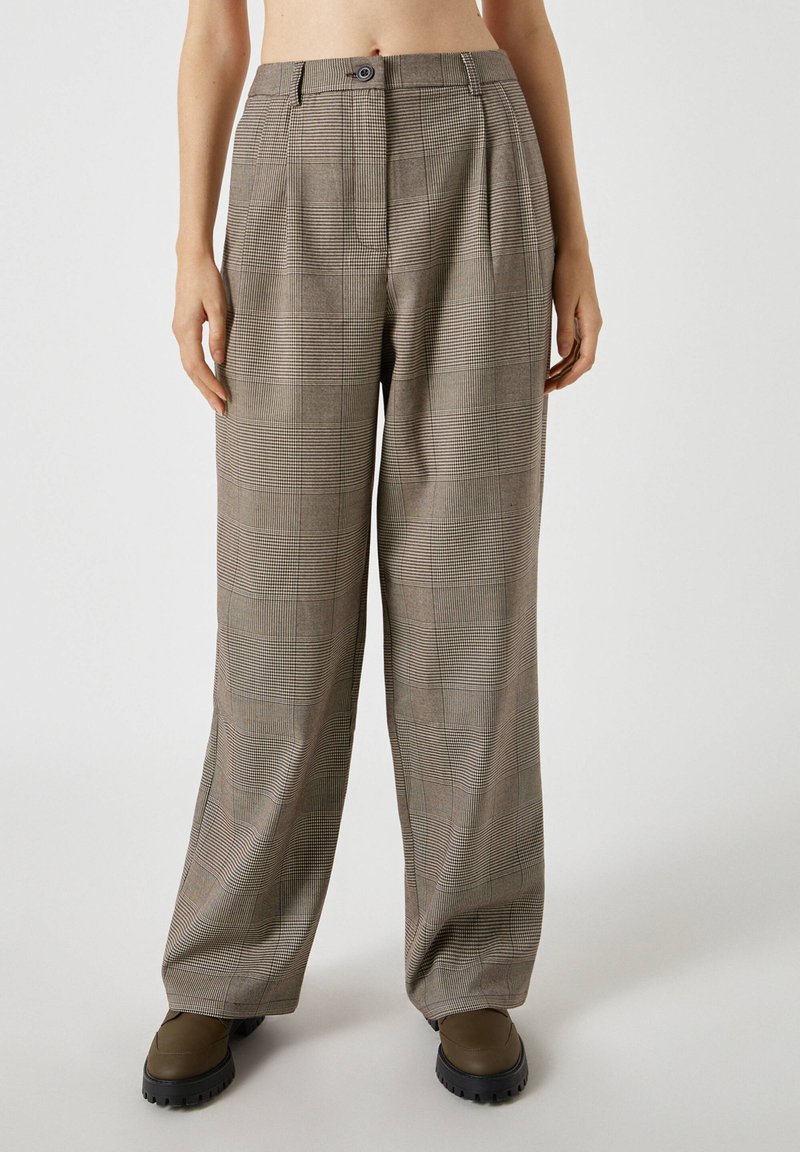 Femme portant un pantalon large à carreaux marron et des chaussures marron épaisses, debout sur un fond clair uni.