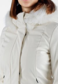Giacca imbottita di colore crema con un morbido collo in finta pelliccia bianco. Presenta tasche con zip e un design snello e aderente con cuciture discrete.