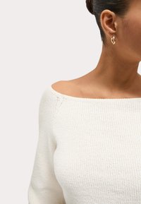 Femme portant un pull en maille blanc à épaules dénudées et des boucles d'oreilles dorées en forme de créole, cheveux attachés, regardant vers la droite sur un fond uni.