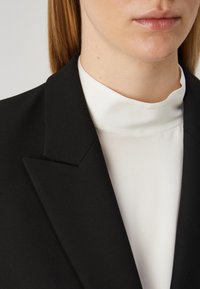 Černý blazer s ostrými klopami, kombinovaný s bílým topem s vysokým výstřihem, vytváří elegantní a strukturovaný vzhled. Hladká textúra látky je viditelná.