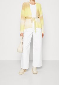 Cárdigan de punto en degradado amarillo y beige, con abertura frontal y cintura ajustada, combinado con pantalones de pierna ancha blancos y accesorios beige.