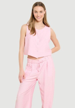 Jauna sieviete smaida, valkā gaiši rozā bezroku pogājamus īsus topiņu un atbilstošas augstu jostas platas bikses, stāvot ar roku uz gurna.