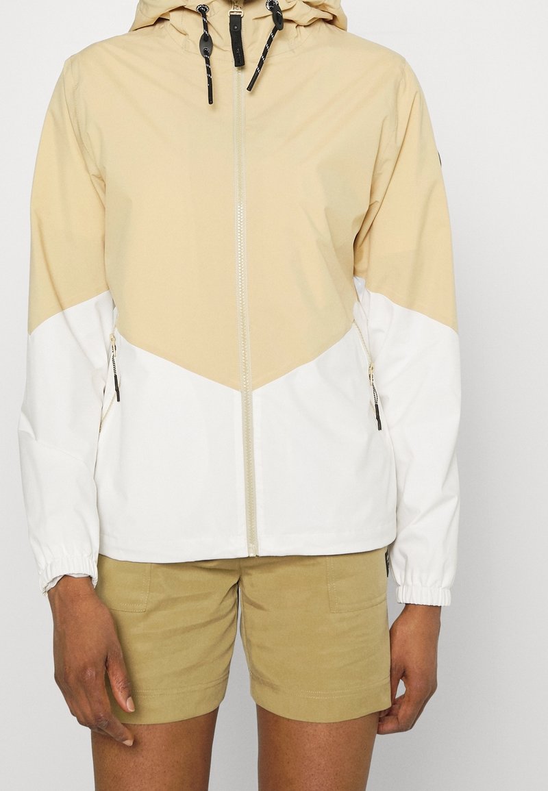 Personne portant une veste à capuche beige et blanche avec fermeture éclair et un short beige, debout devant un fond clair uni.