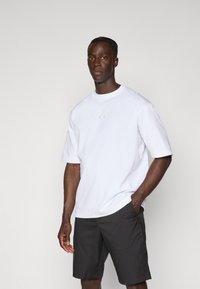 Hvid crew-neck kortærmet t-shirt med et lille logo på brystet, parret med mørke shorts, stående foran en neutral baggrund.