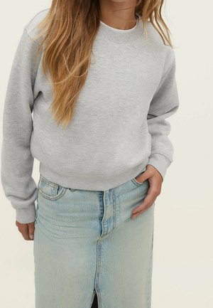 Vrouw met lang haar, gekleed in een lichtgrijze trui en een blauwe denim rok met hoge taille en een voorzijde split, hand rustend in de rokzak.