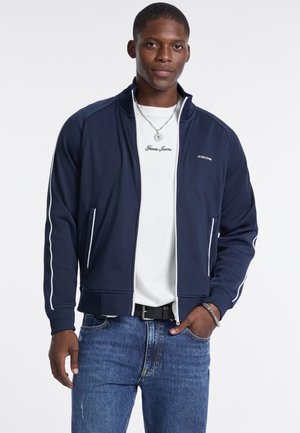 Marineblaue Zip-Jacke mit weißen Streifenakzenten, aus glattem Stoff, getragen über einem weißen T-Shirt. Lässiger Schnitt, mit seitlichen Taschen und gerippten Bündchen.