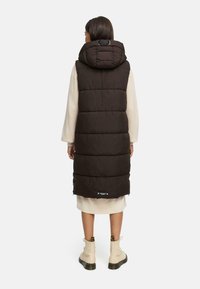 Veste rembourrée marron avec capuche, présentant un design matelassé, un col montant et des poches latérales, portée sur une robe beige et des bottes.
