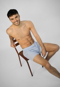 Boxer blu e bianchi a righe con elastico in vita, indossati da un modello seduto. Lo sfondo è grigio uniforme e il modello è a piedi nudi.