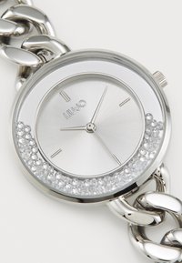 Silver watch with a round face, crystal accents on the bezel, and a chunky metallic chain strap. Displays a textured silver dial.