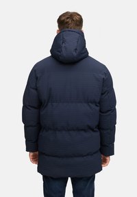 Marinblå pufferjacka med quiltad textur, utrustad med huva, generös vaddering och elastiska ärmslut, designad för värme och komfort.