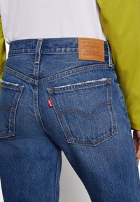Gros plan sur une personne portant un jean en denim bleu Levi's avec des poches arrière classiques et une étiquette en cuir marron de la marque, associée à un chemisier à manches longues blanc et vert.
