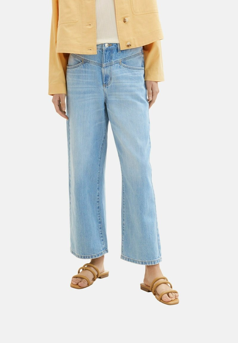Femme portant un jean large bleu clair, des sandales à double bride couleur sable, et une veste jaune par-dessus un haut blanc, debout devant un fond uni.