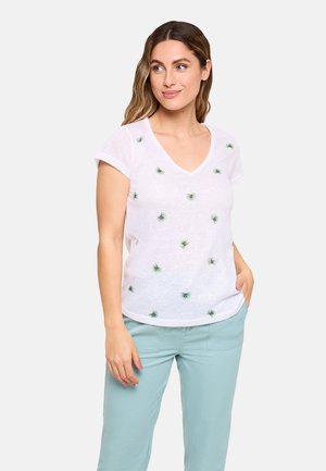 Mujer con una camiseta blanca de cuello en V con pequeños estampados de flores verdes, combinada con pantalones azul claro, de pie con una mano en el bolsillo.