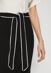 Jupe noire avec une ceinture assortie ornée d'un liseré blanc nouée à la taille, portée avec un haut blanc à manches courtes et un bras droit visible.