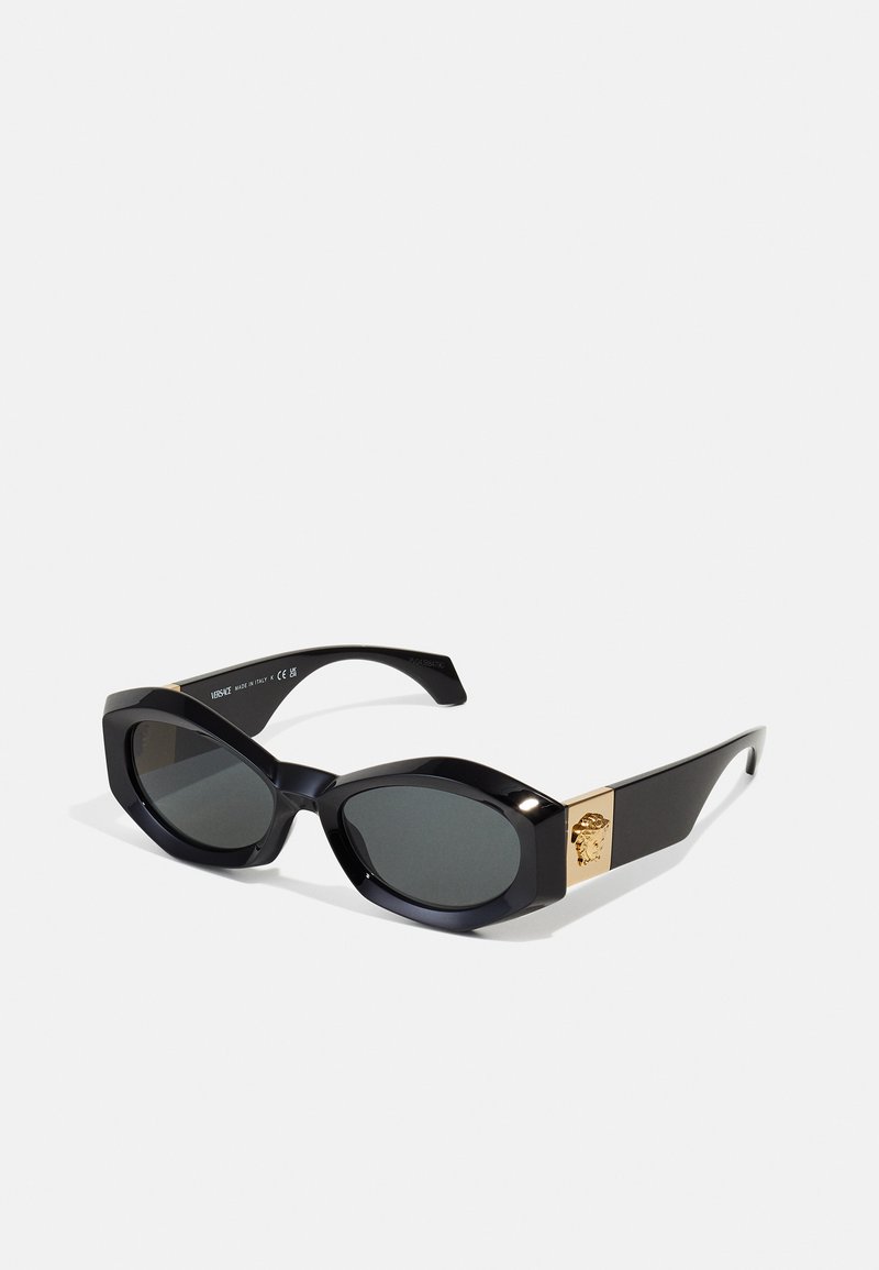 Occhiali da sole neri con un design angolare, montatura in plastica, lenti grigie e un accento con logo dorato sul lato.