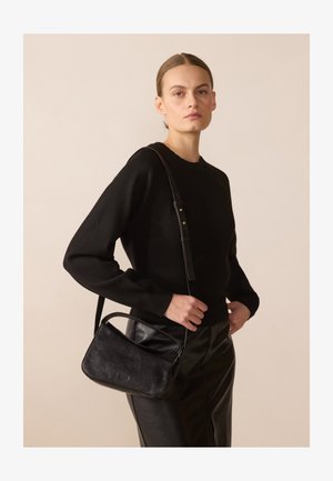 Mustast nahkrist crossbody kott sujuva tekstuuriga, millel on kaarjas kuju, reguleeritav rihm ja reljeefne bränding. Kandestatud musta sviitri kohal.