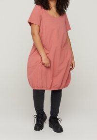 En person med lockigt hår står i en stor rosa klänning och svarta byxor med sneakers, med händerna i fickorna mot en neutral bakgrund.