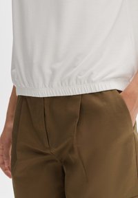Top bianco a maniche corte con orlo elasticizzato abbinato a pantaloni marroni a pieghe con vita alta. Texture lisce, vestibilità rilassata.