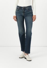 Dark blue denim jeans with a straight-leg cut, five-pocket design, and a slight fade. Paired with white slip-on shoes.