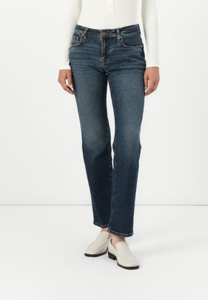 Temno modri jeans z ravnimi hlačnicami, petimi žepi in rahlo bledenjem. Ujemajoči se z belimi copati brez vezalk.