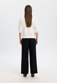 Hvit genser med puffede ermer, kombinert med svarte vidbukser. Glatt stoff og avslappet passform, sett bakfra. Sorte hæler synlige.