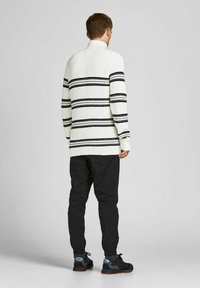 Weißer Strickpullover mit schwarzen horizontalen Streifen, hohem Kragen und Rippstruktur, kombiniert mit schwarzen schmalen Hosen und Sportschuhen.