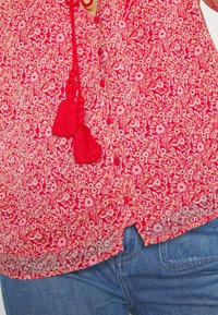 Blouse rouge à motif floral avec boutons et liens à pompons rouges, associée à un jean en denim bleu.