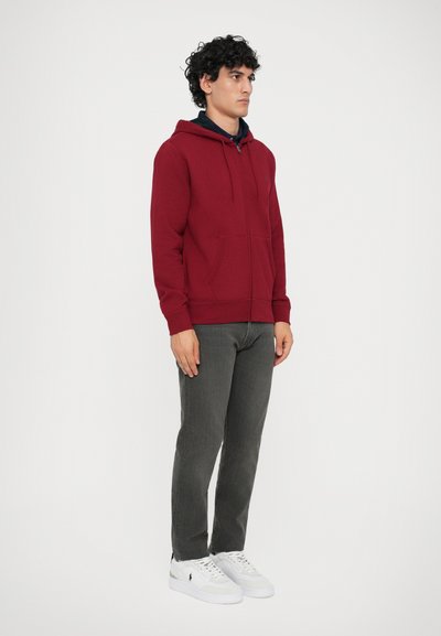Sweatshirt com capuz em cor bordeaux, com bolsos frontais, tecido macio, punhos canelados, sobre uma camisa azul-marinho, combinado com calças cinzentas e ténis brancos.