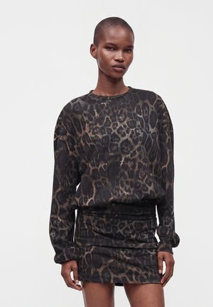 Kvinde iført langærmet leopardprint top og matchende kort nederdel, stående mod en ensfarvet lys baggrund.