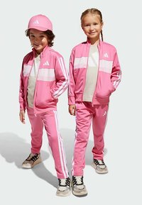 Zwei Kinder tragen abgestimmte, leuchtend pinke Trainingsanzüge aus glattem Material, die weiße Streifen und eine Zip-up-Jacke mit Logoakzenten haben.