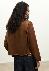 Frau mit langen, welligen Haaren, trägt eine braune Wildlederjacke und dunkelblaue Jeans, steht drinnen vor einem schlichten hellen Hintergrund.