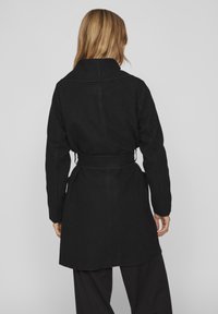 Mustaa vyöllä varustettu takki, jossa on korkea kaulus, valmistettu teksturoidusta kankaasta. Ominaisuuksiin kuuluu pitkät hihat ja polvipituinen leikkaus. Yksinkertainen muotoilu ilman näkyviä kuvioita.