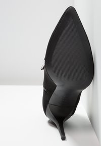 Escarpin en daim noir à talon haut avec un embout pointu, une semelle texturée et une fermeture éclair latérale. Le talon est effilé en forme d'aiguille.
