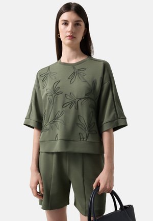 Junge Frau, die ein olivgrünes besticktes Oberteil und passende Shorts trägt, hält eine schwarze Handtasche vor einem einfarbigen Hintergrund.