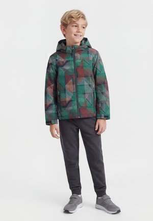 Αγόρι όρθιο και χαμογελαστό, φοράει πολύχρωμο μπουφάν με γεωμετρικά σχέδια και κουκούλα, σκουρόχρωμο παντελόνι και γκρι αθλητικά παπούτσια σε λευκό φόντο.