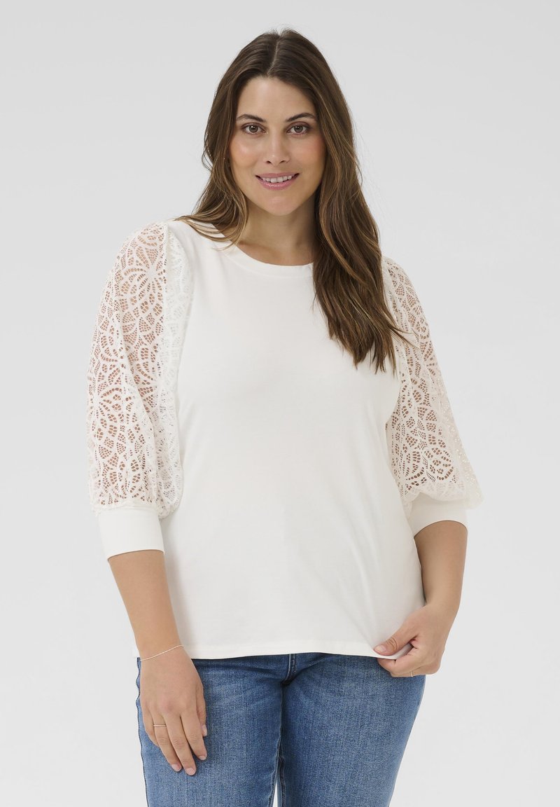 Top blanco de manga larga con detalles intrincados de encaje en las mangas, tela suave, escote redondo y ajuste cómodo. Jeans azules visibles en la parte inferior.