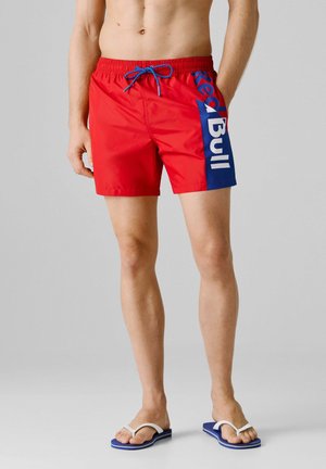 Mann trägt rote Badeshorts mit blauem "Red Bull"-Aufdruck an der Seite und blauem Kordelzug, kombiniert mit blauen und weißen Flip-Flops.