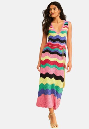 Naine kannab varrukateta mitut värvi laineliste triibudega kudumitest maksikleiti, millel on V-kaelus ja kuldsed kõrge kontsaga sandaalid.