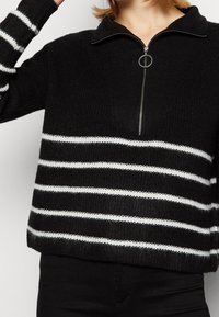 Svart stickad tröja med dragkedja vid kragen, med växlande vita ränder över kroppen och ärmarna, ribbad textur och avslappnad passform.