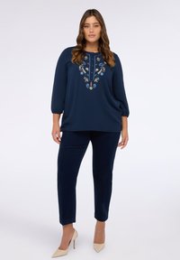 Marineblauwe blouse met driekwartmouwen, versierd met kleurrijke bloemenborduursels, gecombineerd met donkerblauwe broek en beige hoge hakken.